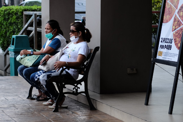 Two women have pulled down the masks covering their faces while sitting on a bench in Dagupan City, Philippines, March 9, 2020. (Jojo Rinoza/BenarNews)