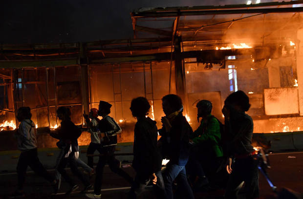 Para pengunjuk rasa melarikan diri dari semprotan gas air mata polisi dengan latar belakang bangunan yang dibakar dalam demonstrasi buruh dan mahasiswa menentang pengesahan Undang-Undang Cipta Kerja yang dinilai mengorbankan hak-hak buruh dan lingkungan hidup, di Jakarta, 8 Oktober 2020. [AFP]